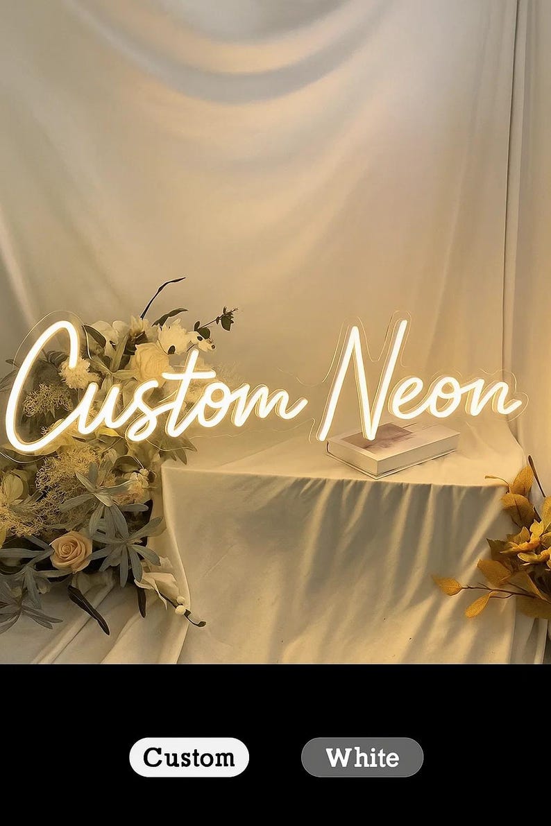 May include: A white neon sign featuring the script text Custom Neon, illuminated with a bright glow. The sign is positioned near a floral arrangement with light-colored flowers and foliage, resting on a draped fabric surface with a book.