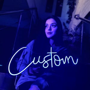 May include: A bright blue neon sign featuring the word Custom in a cursive script font. The sign emits a vibrant glow, illuminating the surrounding area with a cool-toned light. The lettering is fluid and continuous, creating a clear visual display.