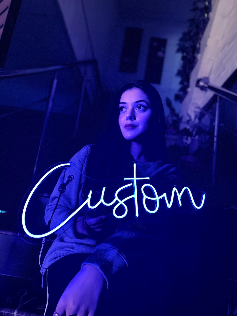 May include: A bright blue neon sign featuring the word Custom in a cursive script font. The sign emits a vibrant glow, illuminating the surrounding area with a cool-toned light. The lettering is fluid and continuous, creating a clear visual display.