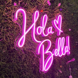 May include: A neon sign with the words "Hola Bella!" in a cursive font, illuminated in bright pink. The sign includes a heart symbol and is mounted on a backdrop of green foliage.