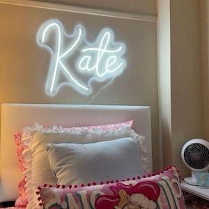 May include: A bedroom scene featuring a white neon sign spelling "Kate" above a bed. The bed has a white headboard, layered pillows, and a pink floral patterned comforter. A decorative pillow with a cartoon character is visible.