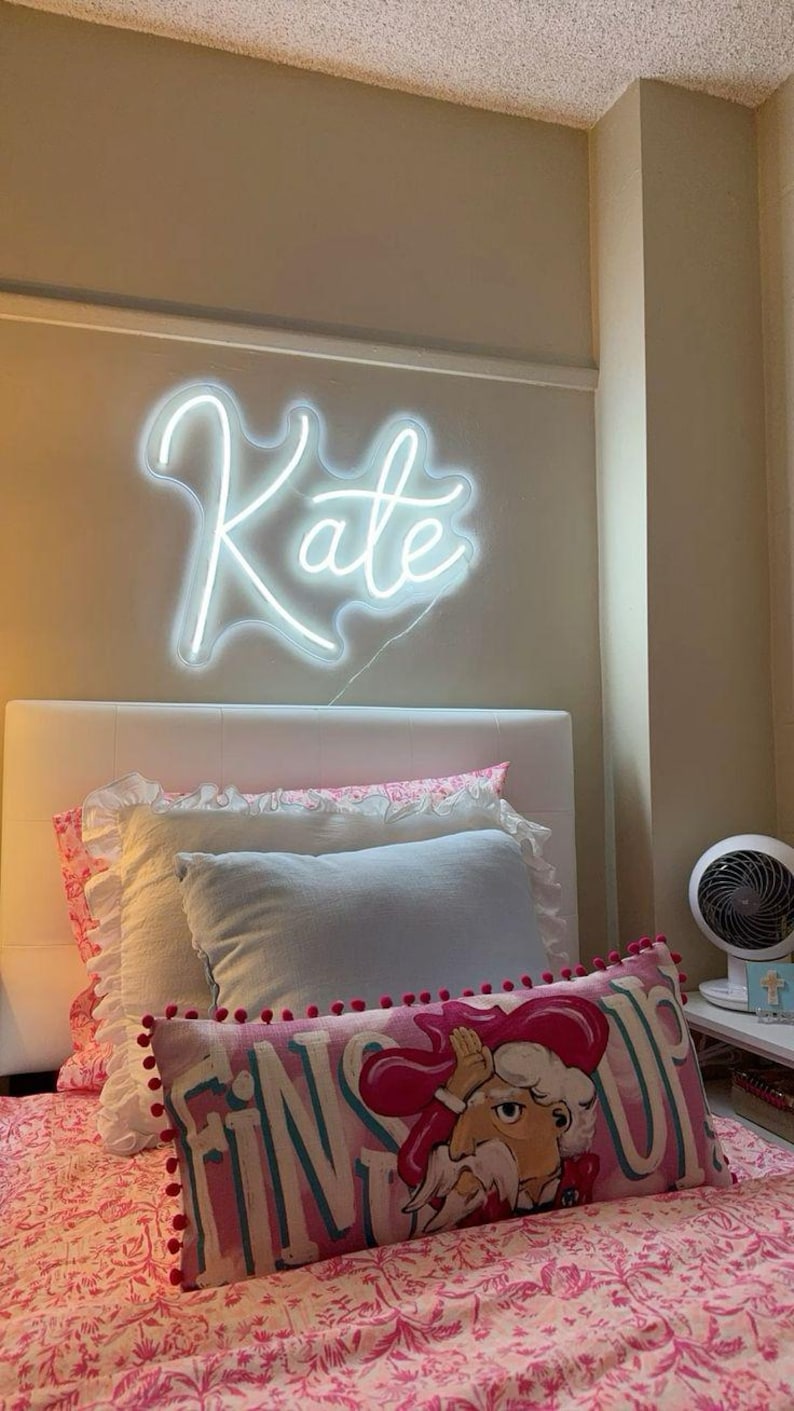May include: A bedroom scene featuring a white neon sign spelling "Kate" above a bed. The bed has a white headboard, layered pillows, and a pink floral patterned comforter. A decorative pillow with a cartoon character is visible.