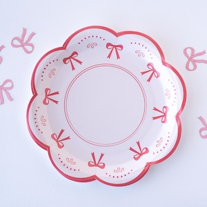 May include: White paper party plate with a scalloped edge and a pink bow design. The plate has a pink border and a pink bow design around the edge.