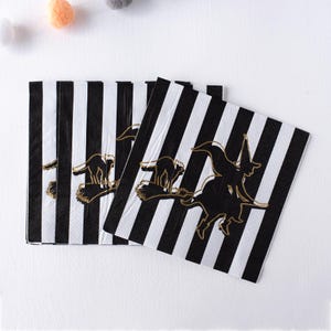 May include: Black and white striped paper napkins with a gold outline of a witch on a broomstick and a black cat. The napkins are folded and stacked, perfect for a Halloween party or spooky event.