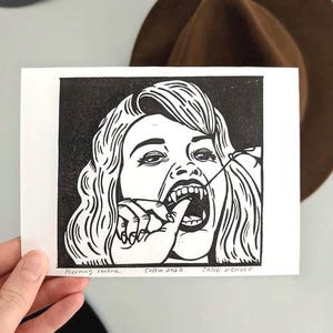 以下が含まれることがあります： 指で唇を引く牙のある人物の様式化された肖像画が描かれた、紙に印刷された白黒の版画。下部には黒インクで「Morning routine」「Sasha 2022」「SANG D'ENCRE」という文字が記されています。
