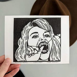 May include: Black and white linocut print of a woman with long wavy hair, pulling at her fang with a string. The print includes the text "Morning routine", "sasha 2022", and "FANG D'ENCRE".