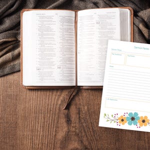 Puede incluir: Una Biblia abierta con una cubierta de cuero marr&oacute;n y un marcador a juego descansa sobre una superficie de madera. Un bloc de notas con las palabras "Notas del serm&oacute;n" y un dise&ntilde;o floral est&aacute; al lado de la Biblia. Una tela marr&oacute;n y gris est&aacute; drapeada en el fondo.