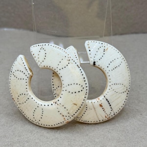 May include: Two off-white, semi-circular earrings with a dotted pattern. The earrings have a unique, open design and are likely made of a natural material. The dotted pattern adds a decorative element to the earrings.