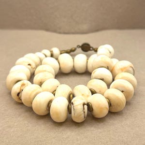 Puede incluir: Collar de cuentas color crema con cierre color bronce. Las cuentas ovaladas tienen un aspecto veteado con marcas marrones y beige. El collar se muestra sobre un fondo neutro.