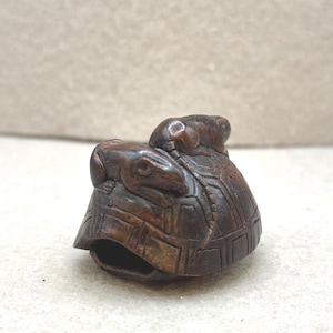 Peut inclure: Une sculpture en bois sculptée à la main représentant une tortue avec une grenouille sur le dos. La carapace de la tortue présente un motif texturé, et la grenouille est perchée sur la tête de la tortue. La sculpture est de couleur marron foncé.