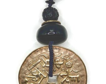 Op de afbeelding: Een vintage bronzen munthanger met een donkerblauw koord en een zwart kralenontwerp. De munt heeft ingewikkelde reliëftekens en symbolen. De hanger heeft een zwarte ring aan de bovenkant.
