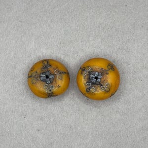 May include: A pair of amber-coloured, circular buttons with detailed silver-toned metal embellishments. Each button has a central square design and delicate swirls. The buttons have a slightly aged look, possibly vintage or antique.