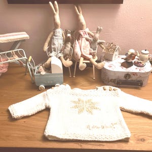 May include: A white knitted sweater with a gold star design and the text "A little bit of magic" on the front. The sweater is laid out on a wooden surface.