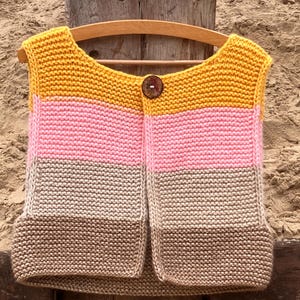 May include: A handmade, knitted vest in a color-blocked design. The vest features horizontal stripes of mustard yellow, pink, and tan. A large, round, brown button fastens the front. The vest is displayed on a wooden hanger.