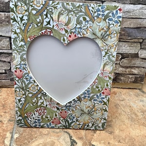May include: A rectangular photo frame with a heart-shaped aperture. The frame features a floral design in green, red, and blue on a white backdrop. The frame is constructed from a durable material.