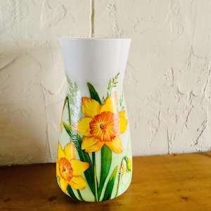 May include: A white ceramic vase with a hand-painted design of yellow daffodils with orange centres and green leaves on a light green background.