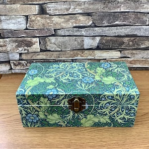 May include: A rectangular wooden box with a green floral pattern and a brass clasp. The box features a repeating design of green leaves and blue flowers on a dark green background. The box is sitting on a wooden surface.