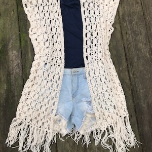 K&ouml;nnte beinhalten: Eine cremefarbene, offene H&auml;kelweste mit Fransen. Die Weste ist &uuml;ber einem marineblauen Oberteil und hellblauen Jeansshorts geschichtet. Das H&auml;keldesign weist ein offenes, spitzenartiges Muster auf. Die Fransen h&auml;ngen am unteren Rand der West.
