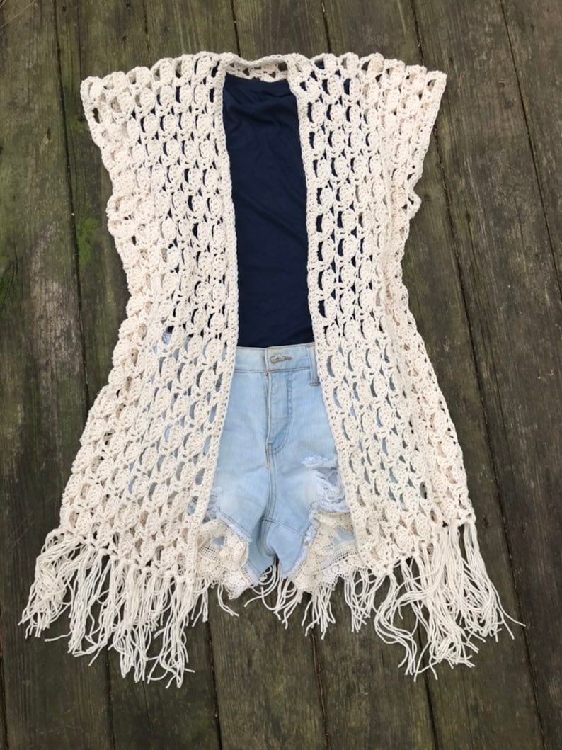 K&ouml;nnte beinhalten: Eine cremefarbene, offene H&auml;kelweste mit Fransen. Die Weste ist &uuml;ber einem marineblauen Oberteil und hellblauen Jeansshorts geschichtet. Das H&auml;keldesign weist ein offenes, spitzenartiges Muster auf. Die Fransen h&auml;ngen am unteren Rand der West.