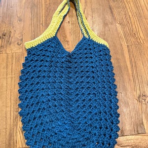May include: A handmade, blue crochet tote bag with yellow handles. The bag features a textured stitch pattern and a V-shaped opening. The handles are a contrasting yellow color, adding a pop of brightness to the design. The bag is likely used for carrying items.