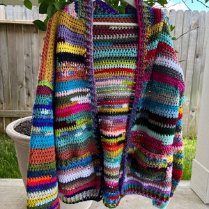 May include: A handmade, open-front crochet cardigan in a vibrant patchwork of colors. The sweater features a mix of horizontal and vertical stripes in shades of blue, green, pink, yellow, and purple. The cardigan is hanging on a white hanger.