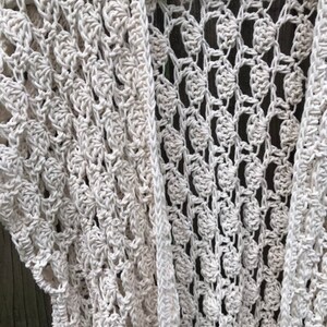 K&ouml;nnte beinhalten: Cremefarbene, offen gewebte H&auml;keljacke. Das Kleidungsst&uuml;ck hat ein zartes, spitzenartiges Muster mit einem strukturierten, bl&uuml;tenartigen Design. Die &Auml;rmel sind kurz und die Strickjacke h&auml;ngt an einem wei&szlig;en Kleiderb&uuml;gel. Der Gesamtstil ist b&ouml;hmisch und handgefertigt.