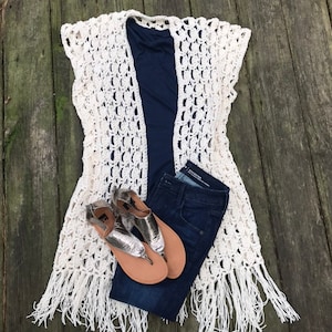K&ouml;nnte beinhalten: Eine cremefarbene, offen gewebte H&auml;kelweste mit Fransen, &uuml;ber einem marineblauen Oberteil und dunkelblauen Jeans. Silberne Sandalen mit beigefarbenen Sohlen vervollst&auml;ndigen das Outfit, das auf einer verwitterten Holzoberfl&auml;che angeordnet ist.