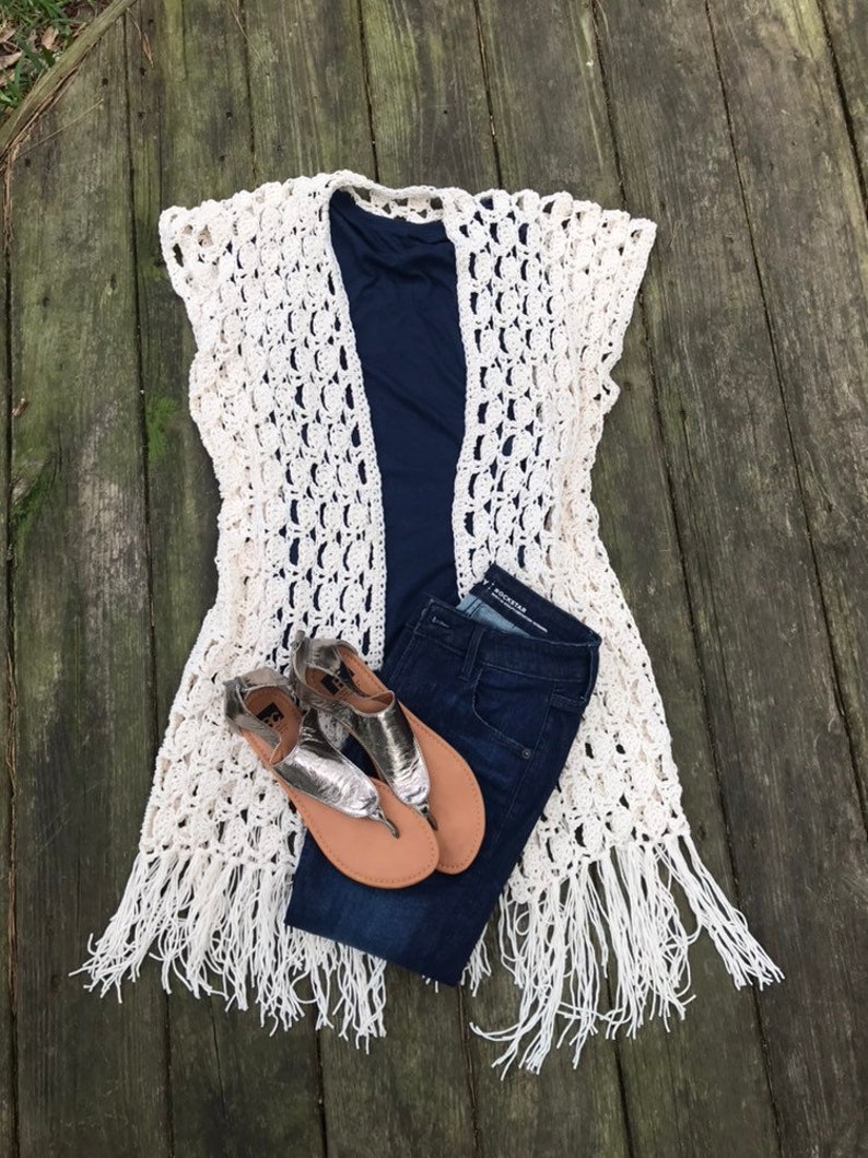 K&ouml;nnte beinhalten: Eine cremefarbene, offen gewebte H&auml;kelweste mit Fransen, &uuml;ber einem marineblauen Oberteil und dunkelblauen Jeans. Silberne Sandalen mit beigefarbenen Sohlen vervollst&auml;ndigen das Outfit, das auf einer verwitterten Holzoberfl&auml;che angeordnet ist.