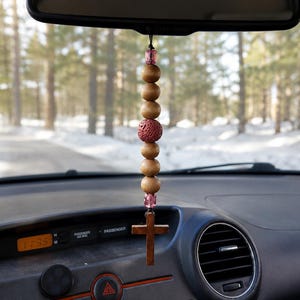 May include: A car rearview mirror hanging ornament featuring a vertical string of wooden beads, two translucent pink faceted beads, and a central porous red lava stone bead, terminating in a small brown wooden cross pendant on a black cord.