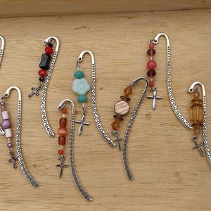 May include: A selection of decorative metal bookmarks with beaded details and cross charms. The bookmarks feature beads in various colours, including blue, red, orange, and brown. The bookmarks are displayed on a light brown wooden surface.