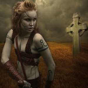 May include: A woman with red hair and a tattoo on her arm stands in a field with a Celtic cross behind her. She is wearing a leather strap across her chest and a leather cuff on her arm. The sky is cloudy and the field is brown.