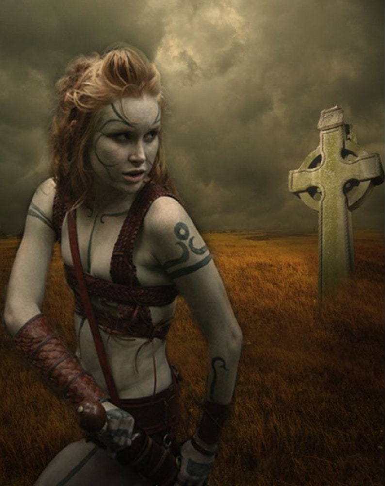 May include: A woman with red hair and a tattoo on her arm stands in a field with a Celtic cross behind her. She is wearing a leather strap across her chest and a leather cuff on her arm. The sky is cloudy and the field is brown.