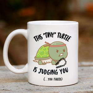 May include: White ceramic mug featuring a cartoon turtle with a green shell and red headband, holding nunchucks. The mug has the text "THIS 'TINY' TURTLE IS JUDGING YOU (...YOU FAILED)".