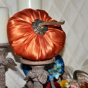 May include: A decorative orange satin pumpkin with a gold metal stem sits on a wooden candlestick. The pumpkin is a fall or autumn themed decoration.
