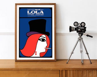 May include: A framed movie poster for Lola, featuring a stylized illustration of a person with bright red hair, a black top hat, and a cigarette. The text reads Lola, Anouk Aimée, and Jacques Demy, set against a deep blue background.