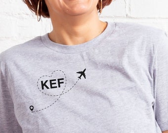 Könnte beinhalten: Eine Frau trägt ein graues T-Shirt mit einer schwarzen Grafik eines Herzens mit den Buchstaben "KEF" darin und einem Flugzeug, das aus dem Herzen fliegt. Das Flugzeug fliegt auf eine Stecknadel auf einer Karte zu.