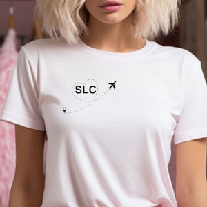 Może przedstawiać: Biała koszulka z czarnym nadrukiem w kształcie serca z tekstem "SLC" w środku, samolotem wylatującym z serca i przerywaną linią prowadzącą do szpilki.