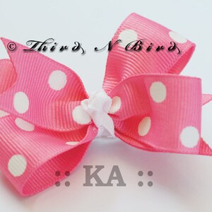 May include: A pink polka dot bow with white spots and a white center knot. The bow is made of a ribbed fabric and has the text "Third N Bird" and "KA" on it.