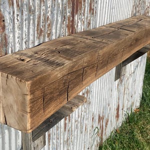 May include: A rectangular wooden beam with a dark brown, weathered finish. The surface displays prominent natural wood grain, deep cracks, and a rough, textured appearance. The beam is mounted horizontally on dark metal support brackets.