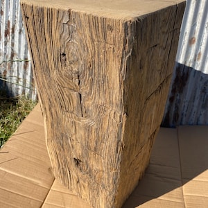 May include: A tall, rectangular block of weathered, light brown wood with a prominent grain pattern, visible knots, and deep vertical fissures. The surface shows a rough, aged texture with natural variations in color and wood grain throughout.