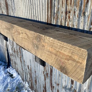 May include: A weathered, rectangular wooden beam, likely a mantel, with a natural, light brown color. The wood grain is visible, and the beam is mounted on a corrugated metal surface. The beam's dimensions are approximately 6 inches high and 8 inches wide.