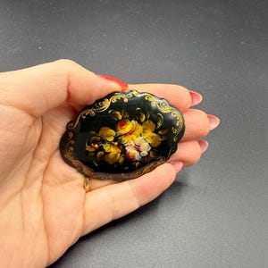 May include: Oval black brooch with a floral design. The brooch features a cluster of yellow, red, and white flowers. The edges are trimmed with gold filigree. The brooch is held in a hand.