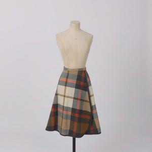 May include: A tartan skirt in brown, beige, red, and grey, shown on a mannequin. The skirt has a classic plaid pattern and a wrap design. The fabric looks like a medium-weight wool or wool blend, suitable for cooler weather. The skirt's length is approximately 60 cm.