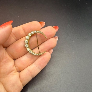 Op de afbeelding: Een broche in de vorm van een halve maan van goudkleurig metaal, voorzien van een rij heldere, rondgeslepen strass-steentjes langs de gebogen rand. Het sieraad is aan de achterzijde voorzien van een dunne, rechte metalen speld.