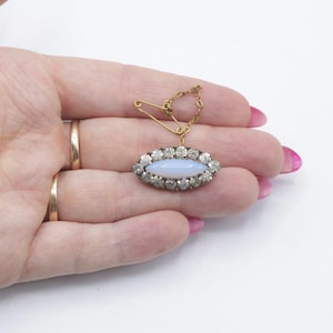 May include: An oval-shaped brooch featuring a central light blue cabochon stone surrounded by a halo of clear rhinestones. The piece is attached to a gold-toned safety pin by a delicate metal chain, creating a dangling pendant effect.