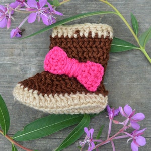 May include: Brown and beige crocheted baby booties with a pink bow on top. The booties are on a wooden surface with purple flowers.