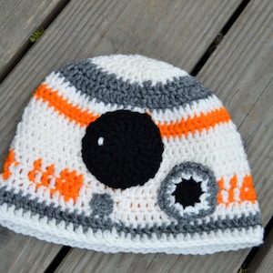 Może przedstawiać: Biały, pomarańczowy i szary kapelusz na szydełku z czarno-białym wzorem przypominającym postać z Gwiezdnych Wojen.