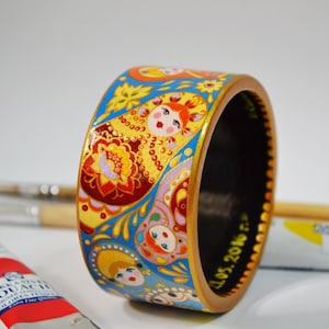 Op de afbeelding: Een handbeschilderde houten armband met een ontwerp van Russische matroesjka poppen. De armband is voornamelijk blauw, geel en rood, met gouden accenten. De binnenkant is zwart en de datum "11.05.2016" staat aan de binnenkant. Diameter: ongeveer 6 cm.
