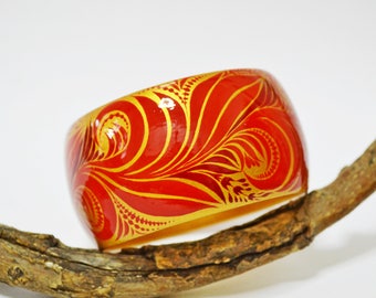 May include: A red and gold wooden bangle with a floral design. The bangle is resting on a branch.