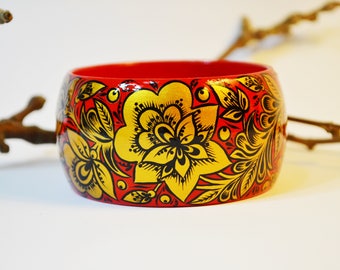 May include: A red wooden bangle with a gold and black floral design. The bangle is painted with a traditional Russian folk art style.